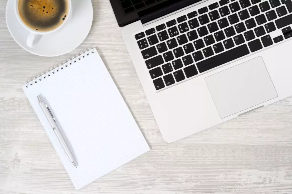 Un laptop, un piccolo taccuino con una matita appoggiata sopra e una tazza di caffè bianca in ceramica con un cucchiaino dentro su un tavolo di legno.