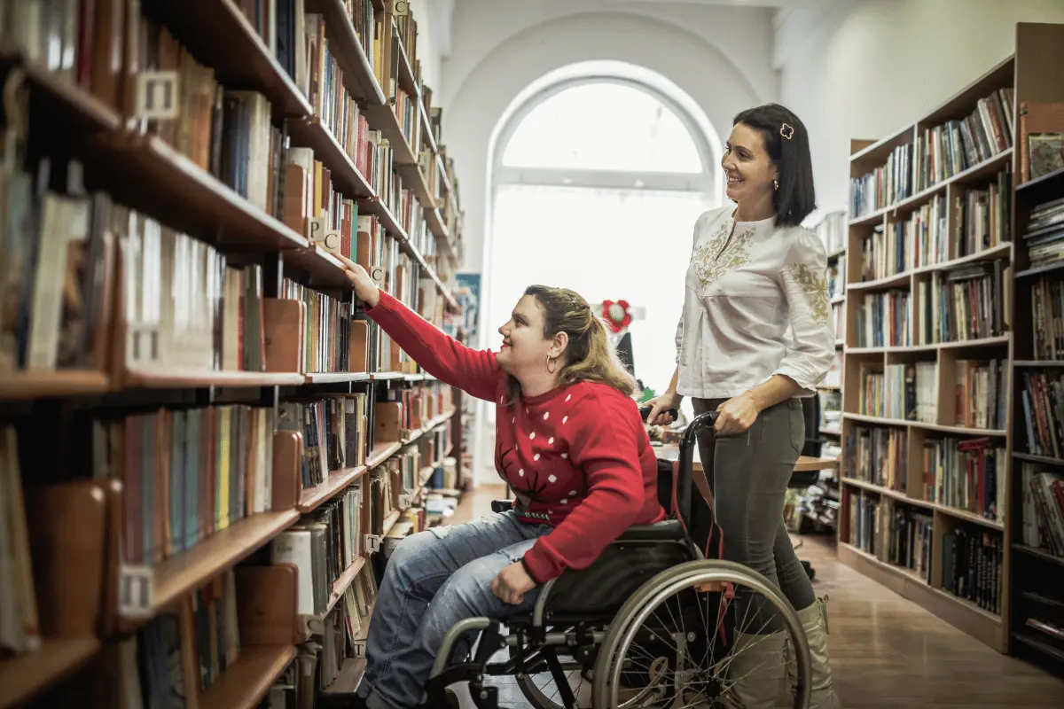 alunna sulla sedia a rotelle nella biblioteca scolastica assistita da un'Asacom