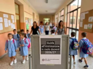 Immissioni in ruolo docenti 2026/27: Docenti e alunni nel corridoio di una scuola. In primo piano la scritta: "Guida all'immissione in ruolo docenti 2026/27"